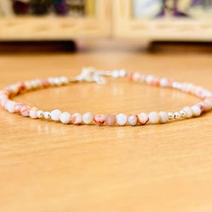 May include: A delicate bracelet composed of small, faceted, multi-colored stone beads in shades of cream, peach, and soft pink, interspersed with tiny, polished silver-toned metallic beads, featuring a silver-toned metal clasp closure.