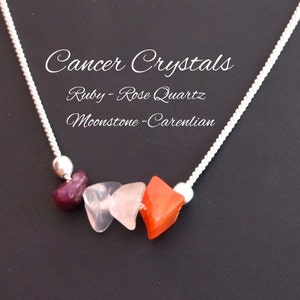 May include: Silver chain necklace with four gemstone beads. The beads are red, pink, and orange. The text "Cancer Crystals" is above the beads. The text "Ruby - Rose Quartz" is below the beads. The text "Moonstone - Carelian" is below the text "Ruby - Rose Quartz".