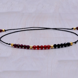 Puede incluir: Un brazalete de cuerda negra con cuentas de piedras preciosas rojas, naranjas y negras.