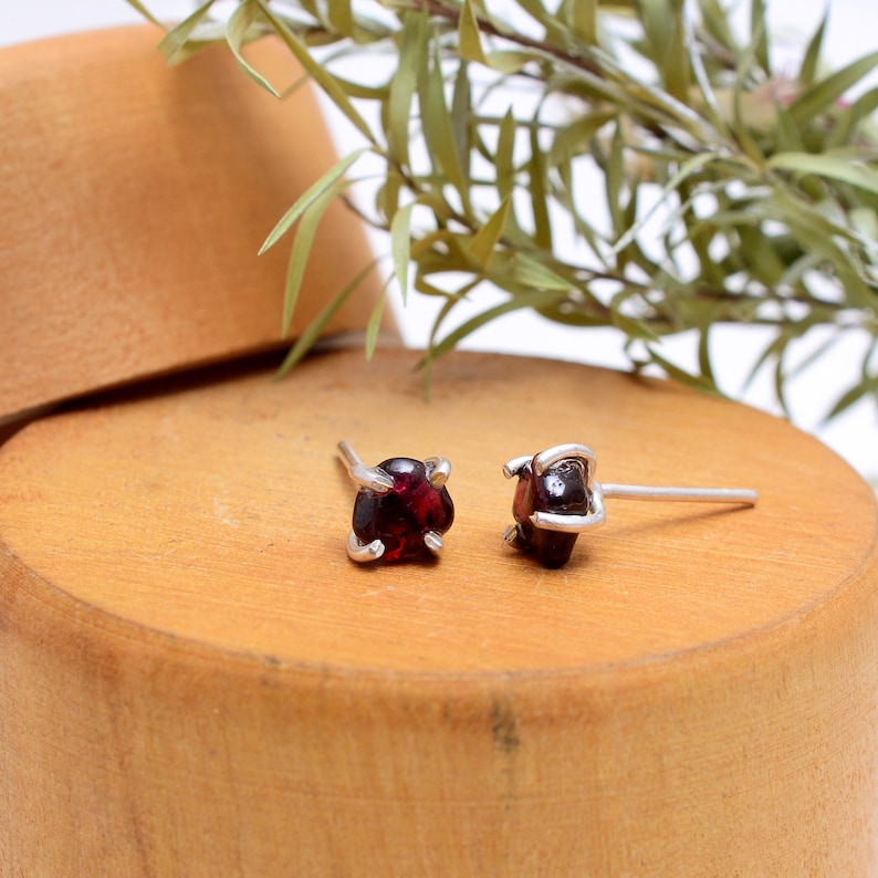 May include: A pair of stud earrings featuring square-cut, deep red gemstones set in silver-colored metal. The earrings are displayed on a wooden surface with a natural wood grain, with green foliage in the background.