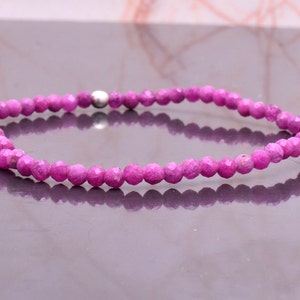 Puede incluir: Una pulsera de cuentas circular compuesta por numerosas cuentas pequeñas, facetadas y de color magenta vibrante. La hebra presenta una única cuenta metálica lisa de tono plateado situada cerca de la parte superior, que contrasta con el acabado mate y texturizado de las cuentas magenta circundantes.
