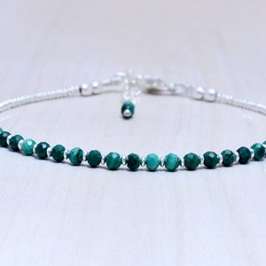 Può includere: Un delicato braccialetto con perline sfaccettate di malachite verde scuro infilate su una catena d'argento. Il braccialetto ha una piccola perla pendente e una chiusura sicura. Il gioiello è esposto su una superficie colorata chiara.