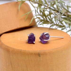 May include: A pair of amethyst stud earrings. Each earring features a raw, deep purple amethyst crystal held in a silver-colored setting. The earrings are displayed on a wooden surface, with a blurred background.
