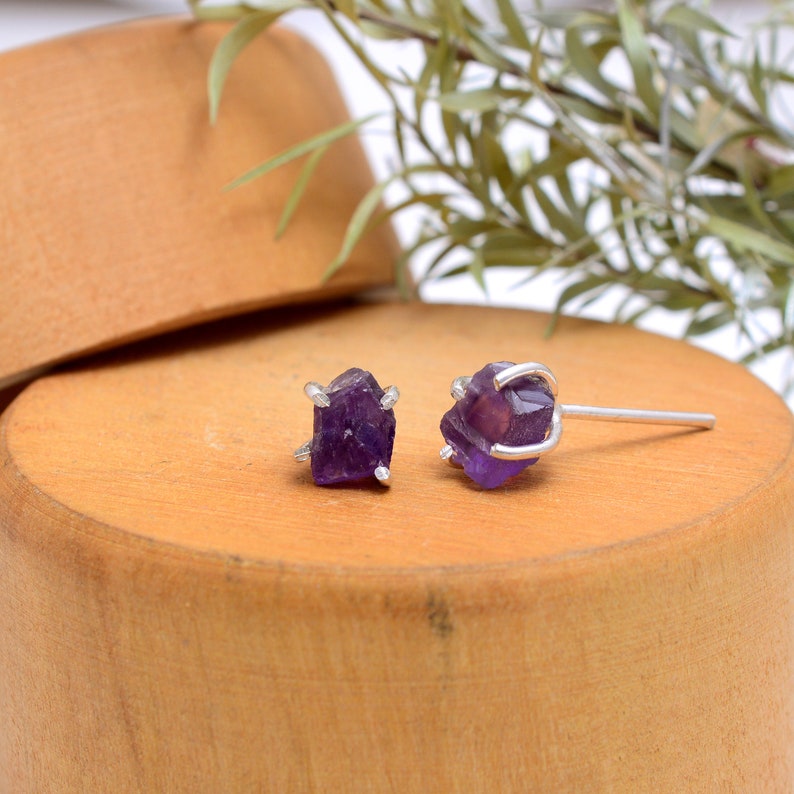 May include: A pair of amethyst stud earrings. Each earring features a raw, deep purple amethyst crystal held in a silver-colored setting. The earrings are displayed on a wooden surface, with a blurred background.