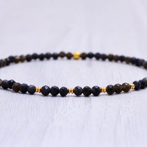 Könnte beinhalten: Ein Armband mit kleinen, facettierten schwarzen Perlen mit grün-goldenen Akzenten. Das Armband ist mit goldfarbenen Perlen und einer einzelnen größeren goldenen Perle verziert. Der Schmuck wird auf einer hellen Holzoberfläche präsentiert.