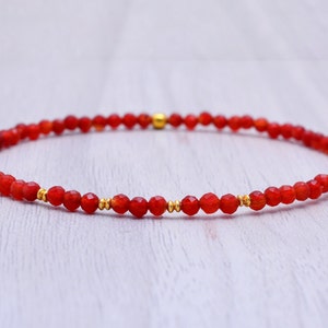 Könnte beinhalten: Ein rotes Karneol-Armband mit goldenen Perlen. Das Armband besteht aus kleinen, facettierten Perlen und ist auf einer elastischen Schnur aufgefädelt.