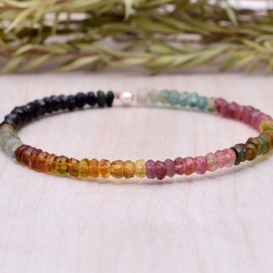 Peut inclure: Un bracelet de perles multicolores, allant du noir au rose. Le bracelet est composé de petites pierres précieuses facettées. Une petite perle blanche est visible au centre du bracelet.