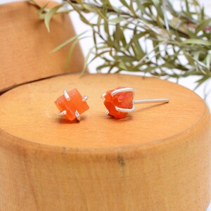 May include: A pair of orange carnelian stud earrings. Each earring features a raw, cube-shaped carnelian stone held in place by a silver metal setting. The earrings are displayed on a wooden surface, with greenery in the background.