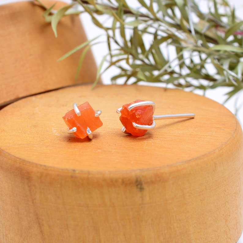 May include: A pair of orange carnelian stud earrings. Each earring features a raw, cube-shaped carnelian stone held in place by a silver metal setting. The earrings are displayed on a wooden surface, with greenery in the background.
