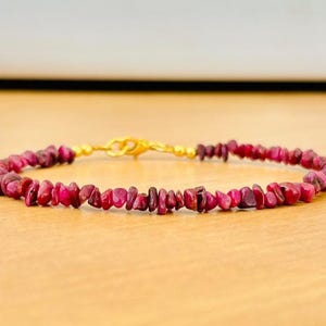 May include: A delicate bracelet featuring small, irregularly shaped, deep red gemstones. The gemstones are strung together with a gold-colored chain and clasp. The bracelet is displayed on a light brown surface.