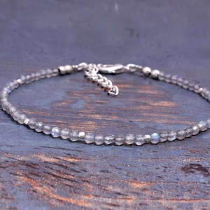 May include: A delicate bracelet featuring small, faceted, grey beads strung together. The bracelet has a silver-coloured chain and clasp. The beads appear to be made of a translucent material, with subtle hints of blue and grey.