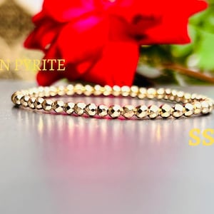 Puede incluir: Un brazalete de cuentas de pirita dorada con el texto "GOLDEN PYRITE" y "SSJ" sobre una superficie blanca.