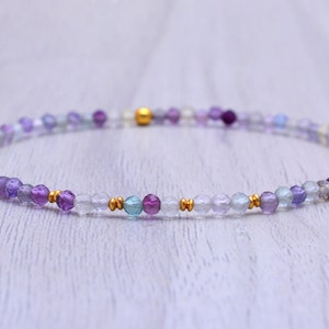 Può includere: Un braccialetto delicato realizzato con piccole perline rotonde in tonalità di viola, blu e trasparente. Le perline sono intervallate da minuscole perline dorate.