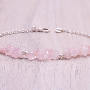 May include: A delicate silver chain bracelet featuring a row of pale pink rose quartz chips. The bracelet is displayed against a light wood background.