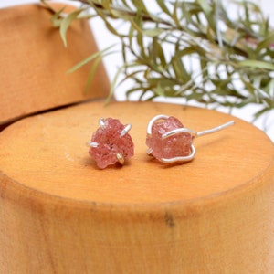 May include: A pair of raw, pink gemstone stud earrings. Each earring features a rough-cut, irregular-shaped pink stone held in place by a silver-colored metal wire and post. The earrings are displayed on a wooden surface.