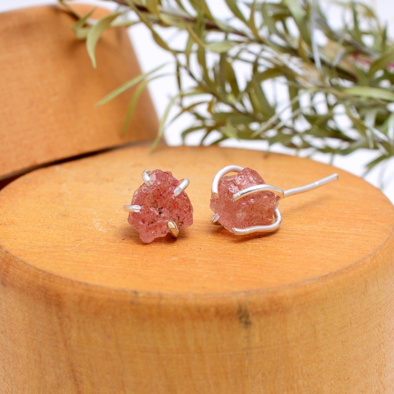 May include: A pair of raw, pink gemstone stud earrings. Each earring features a rough-cut, irregular-shaped pink stone held in place by a silver-colored metal wire and post. The earrings are displayed on a wooden surface.