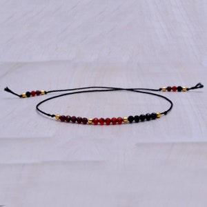 May include: A delicate black string bracelet with red and black beads. The bracelet features a mix of red and black beads, with gold accents.