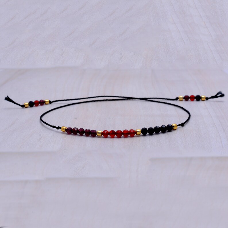 May include: A delicate black string bracelet with red and black beads. The bracelet features a mix of red and black beads, with gold accents.