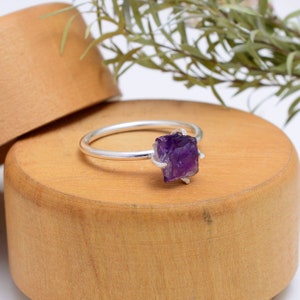 May include: A silver ring featuring a raw amethyst gemstone. The amethyst is a deep purple colour and has a natural, rough texture. The ring is displayed on a wooden surface.