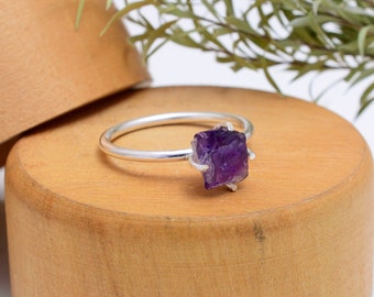 May include: A silver ring with a raw amethyst gemstone. The amethyst is a deep purple color and has a natural, rough texture. The ring is set on a wooden surface.