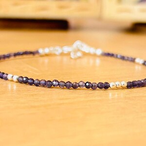 May include: Delicate beaded bracelet featuring small, faceted purple beads interspersed with silver-colored beads. The bracelet is strung on a thin, flexible cord and has a silver-colored clasp. The jewelry is displayed on a light brown surface.