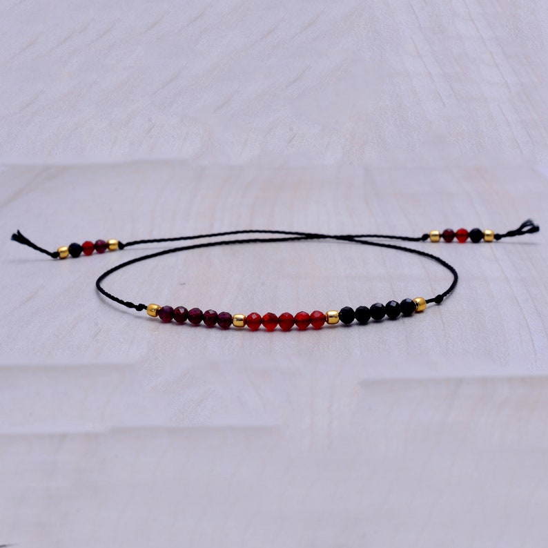 May include: A delicate black string bracelet with red and black beads. The bracelet features a mix of red and black beads, with gold accents.