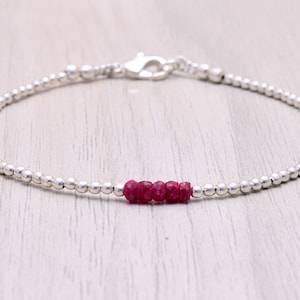 Puede incluir: Una delicada pulsera de cuentas plateadas con cierre de mosquetón. La pulsera presenta cinco piedras preciosas de rubí rojo intenso. La pulsera se muestra sobre una superficie de vetas de madera clara.