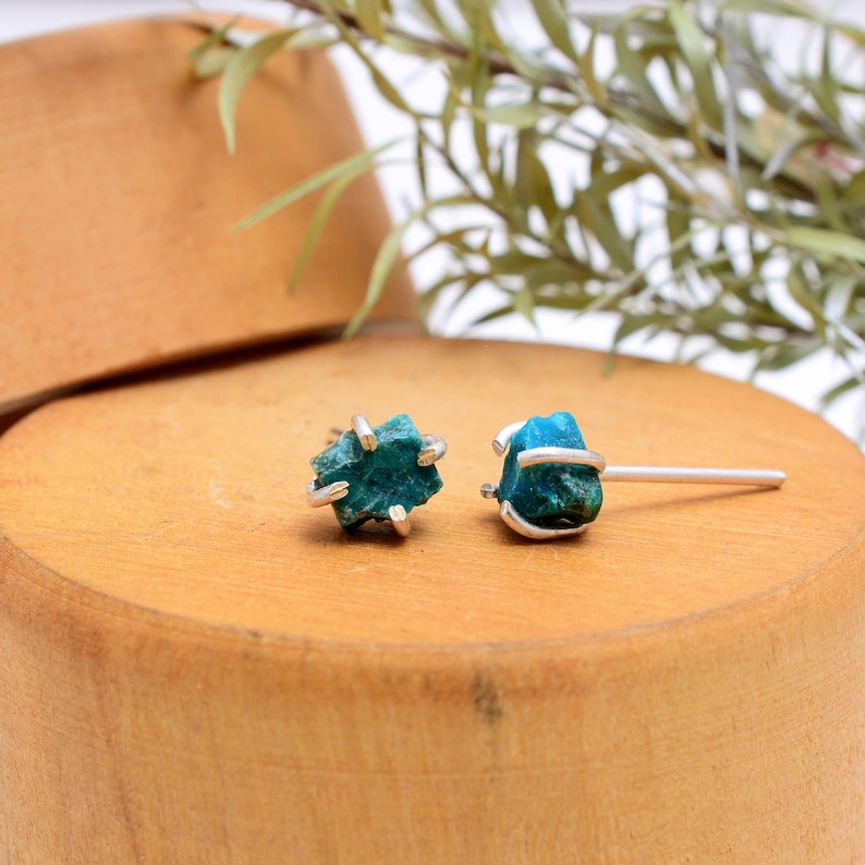 May include: A pair of teal-colored raw stone stud earrings with silver-colored metal settings. The earrings are displayed on a wooden surface, with a blurred background of greenery.