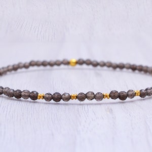 May include: A delicate bracelet featuring small, faceted smoky quartz beads. The beads are strung together with gold-colored accents. This minimalist jewelry piece is designed for everyday wear.