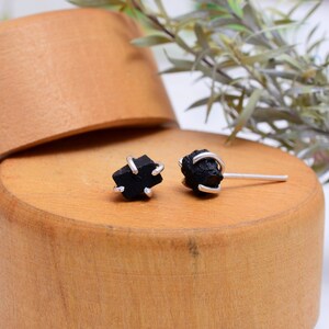 May include: A pair of black tourmaline stud earrings. Each earring features a raw, dark gemstone held in place by a silver-colored metal setting. The earrings are displayed on a wooden surface.