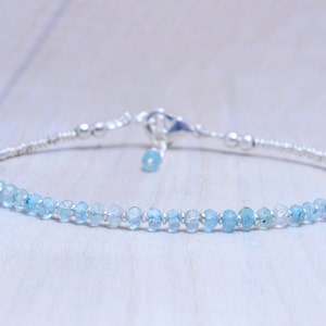 May include: Delicate bracelet featuring small, light blue aquamarine gemstones strung on a thin, silver chain. The bracelet has a secure clasp and a small aquamarine charm. A simple and elegant piece of jewelry.