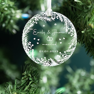 K&ouml;nnte beinhalten: Ein transparenter Weihnachtsbaumschmuck aus Glas mit einem wei&szlig;en Design aus einem Kranz, den Worten "Emily & John married 01.01.2023" und zwei Ringen.