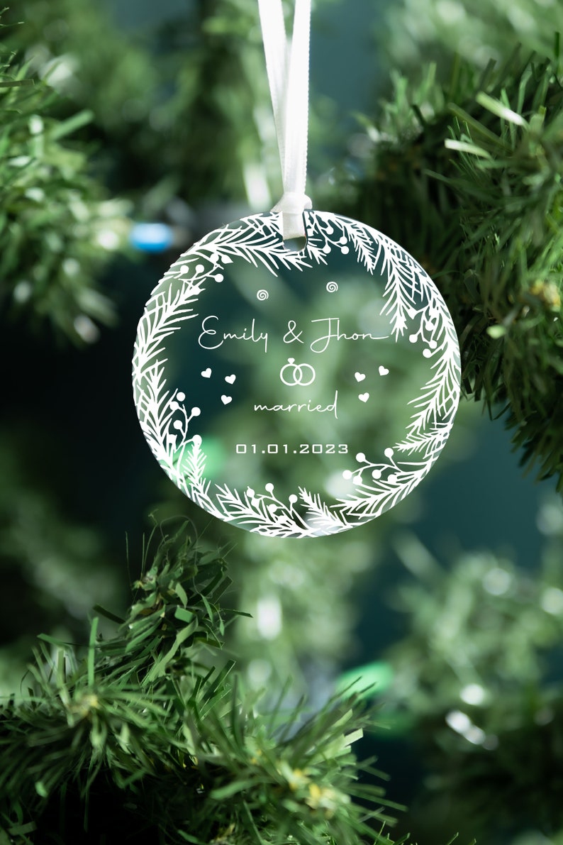 K&ouml;nnte beinhalten: Ein transparenter Weihnachtsbaumschmuck aus Glas mit einem wei&szlig;en Design aus einem Kranz, den Worten "Emily & John married 01.01.2023" und zwei Ringen.