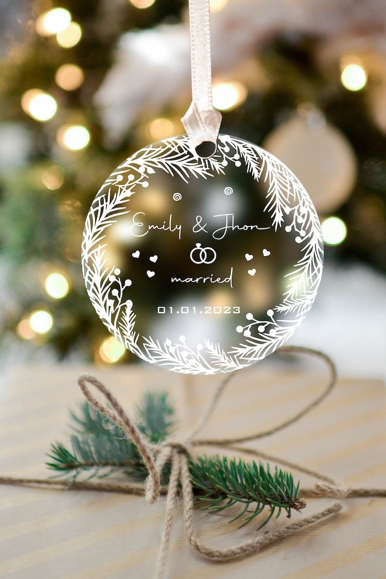 K&ouml;nnte beinhalten: Ein transparenter Weihnachtsbaumschmuck aus Glas mit einem wei&szlig;en Kranzdesign. Der Schmuck zeigt den Text "Emily & John married 01.01.2023" und zwei Ringe.