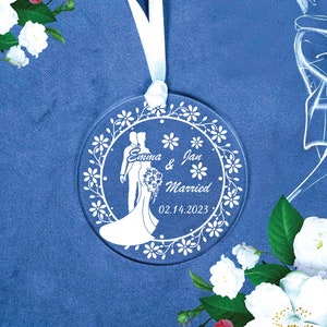 May include: White glass ornament with a floral design and a silhouette of a bride and groom. The text reads "Emma & Jan Married 02.14.2023".