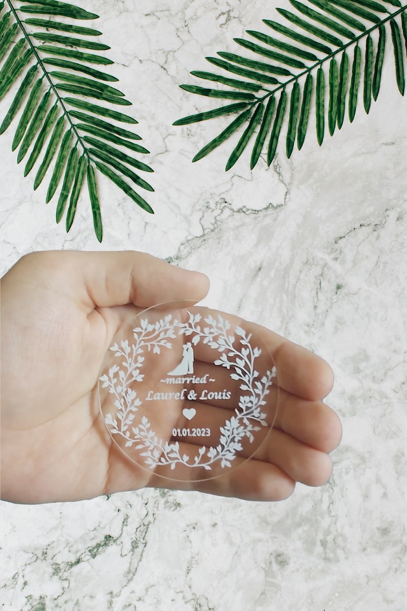 K&ouml;nnte beinhalten: Ein runder, transparenter Acryl-Hochzeitsgeschenkanh&auml;nger mit wei&szlig;em Design. Das Design zeigt ein Paar, ein Herz und den Text "~married~ Laurel & Louis 01.01.2023".