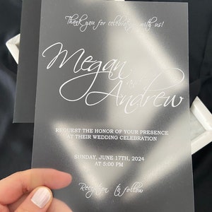 Puede incluir: Una invitación de boda de acrílico transparente con un fondo esmerilado. La invitación presenta los nombres "Megan y Andrew" en una fuente cursiva blanca. El texto "¡Gracias por celebrar con nosotros!" está en la parte superior de la invitación. La fecha y la hora de la boda se enumeran debajo de los nombres. El texto "Recepción a continuación" está en la parte inferior de la invitación.