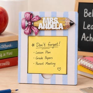May include: A rectangular white and light blue striped wooden note holder featuring a decorative pencil graphic with a pink bow and the text MRS. ANDELA. A yellow sticky note is attached with the handwritten text Don't forget! Lesson Plan, Grade Papers, Parent Meeting.