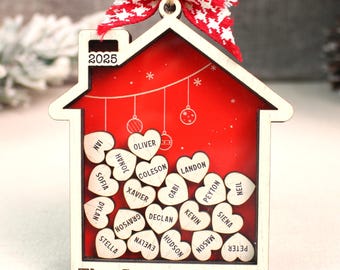 Könnte beinhalten: Weihnachtsschmuck in Form eines Holzhauses mit roter Innenausstattung und dem Text "2025". Der Schmuck hat eine rot-weiße Schleife, Holzherzen mit Namen und den Text "The Smiths Family".