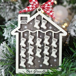 May include: Wooden Christmas ornament in the shape of a house, with the year 2025. The ornament has a dark wood background with white stockings, each with a name. A red and white bow is at the top. The text "Our Grandkids" is at the bottom.