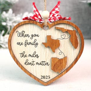 May include: Wooden heart-shaped ornament with the text "When you are family the miles don't matter" and the year 2025. The ornament features cutouts of the states of Texas, Florida, and Ohio, connected by dotted lines. It has a red and white bow.