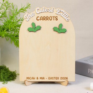 May include: A wooden Easter-themed sign with the words "The Cutest Little Carrots" in a decorative font. The sign has green carrot top accents and the names "Micah & Mia - Easter 2026" at the bottom. The sign is supported by a wooden stand.