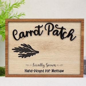 May include: A wooden framed sign with the words "Carrot Patch" in a black, cursive font. Below the text is a black illustration of a leafy sprig. The sign reads "Locally Grown, Hand-Picked for Memaw."