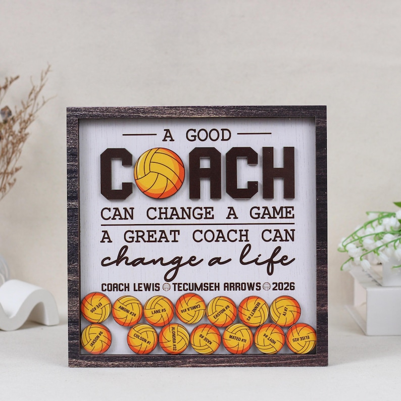 May include: A square wooden shadow box frame containing a white background with the text A GOOD COACH CAN CHANGE A GAME A GREAT COACH CAN CHANGE A LIFE. Below the text are small wooden volleyballs, each personalized with a name and number.