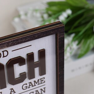 May include: A rectangular wooden sign with a dark brown frame and a white background. The sign features raised, dark brown, three-dimensional lettering that reads "OD" and "CH" with "A GAME" and "CAN" visible below. The wood grain is visible on the frame.