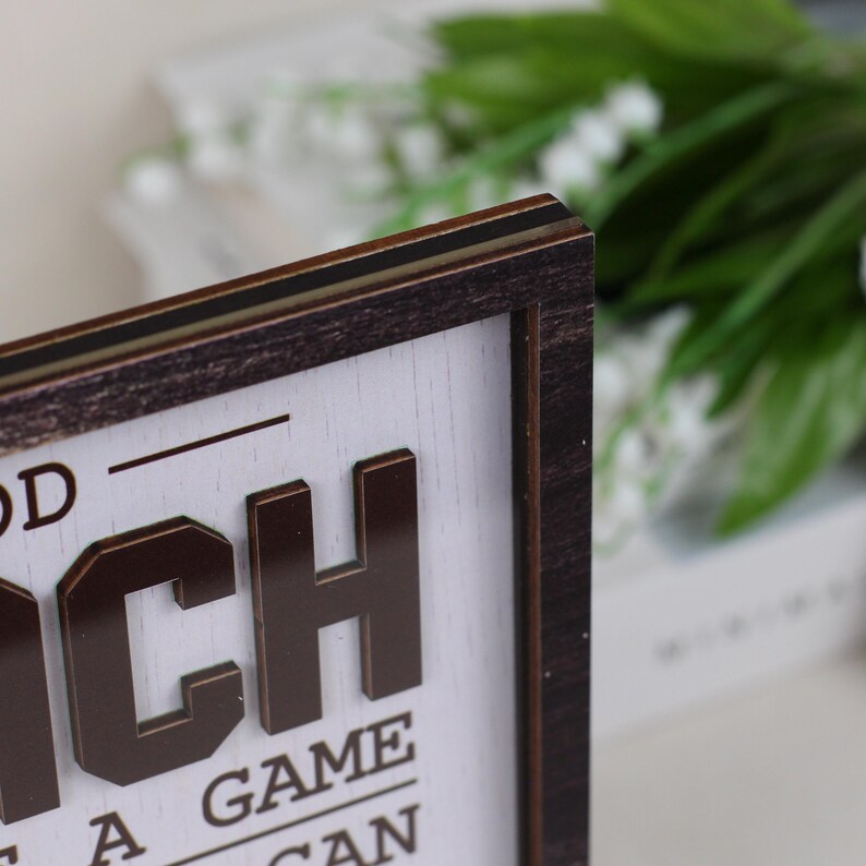 May include: A rectangular wooden sign with a dark brown frame and a white background. The sign features raised, dark brown, three-dimensional lettering that reads "OD" and "CH" with "A GAME" and "CAN" visible below. The wood grain is visible on the frame.