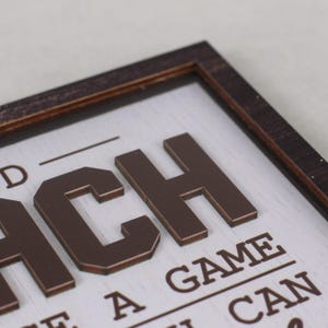 May include: A rectangular wooden sign with a dark brown frame and a white background. The sign features raised, dark brown, block-style lettering that spells out the word COACH, with additional text below reading A GAME, HE, A CAN.