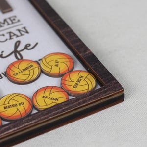 May include: A dark wood-framed shadow box containing circular wooden tokens printed with yellow and orange volleyball graphics. Each token features a name and number, including BENNETT #3, AJ #7, ELISE #25, HOYT #4, and MATEO #11, set against a white background.