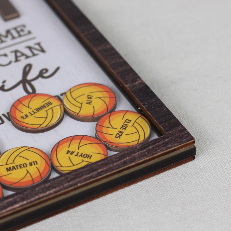 May include: A dark wood-framed shadow box containing circular wooden tokens printed with yellow and orange volleyball graphics. Each token features a name and number, including BENNETT #3, AJ #7, ELISE #25, HOYT #4, and MATEO #11, set against a white background.
