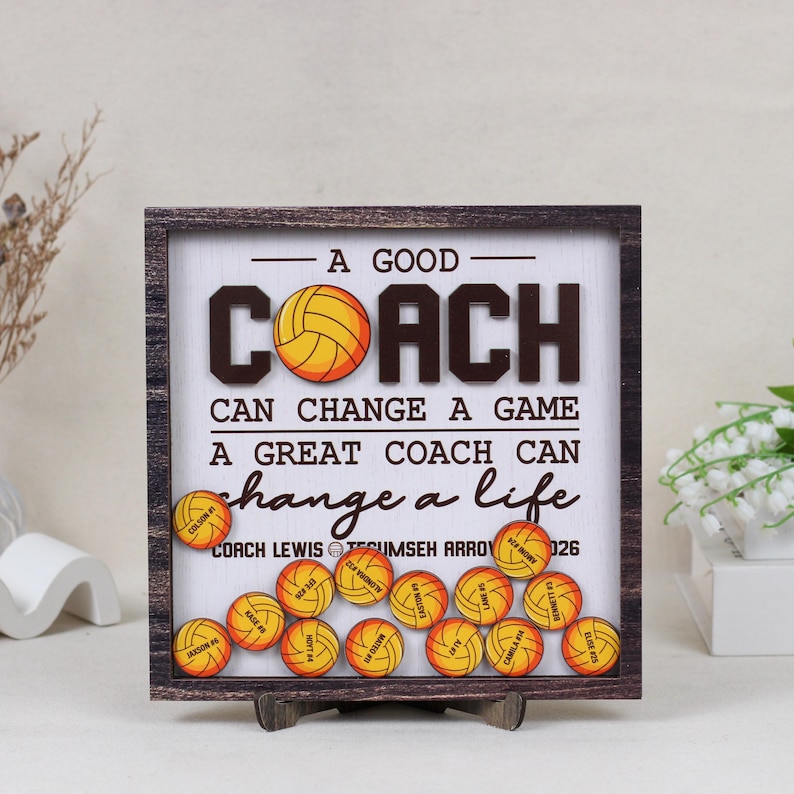 May include: A square wooden shadow box frame containing a white background with the text A GOOD COACH CAN CHANGE A GAME A GREAT COACH CAN CHANGE A LIFE. It features small orange volleyball-shaped tokens with names and numbers printed on them.